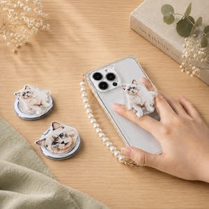 May include: A clear smartphone case with a white pearl beaded wrist strap and a cat-shaped acrylic phone grip. Two additional cat-themed acrylic phone grips are displayed separately, each featuring a detailed illustration of a fluffy cat.