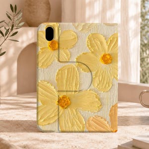 Könnte beinhalten: Eine rechteckige Tablet-Hülle mit einem strukturierten Gemälde im Impasto-Stil, das gelbe Blumen mit leuchtend orangefarbenen Mitten auf cremefarbenem Hintergrund zeigt. Das Design enthält eine Kameraaussparung oben links.