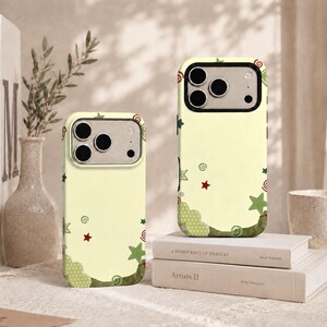 May include: Two pale yellow smartphone cases featuring a whimsical pattern of small red stars, green stars, green polka-dot clouds, and green spiral swirls. The cases have a black camera cutout frame and are displayed in an upright position.