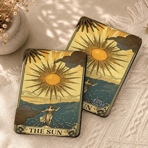 May include: Two rectangular coasters featuring a tarot card illustration titled THE SUN. The design depicts a radiant yellow sun with sharp rays above a landscape with a figure, dark clouds, and blue hills. One coaster includes the custom text EM.