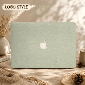 May include: A sage green laptop cover with a textured, faux-leather finish and a central cutout revealing the Apple logo. The rectangular protective case features a consistent, fine-grained pattern across its entire surface, designed to fit a laptop.