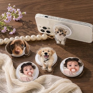 May include: A clear smartphone case with a circular kitten-shaped grip attached, alongside three circular phone grips featuring a brown dachshund, a golden retriever puppy, and a baby with a pacifier, all arranged on a wooden surface.