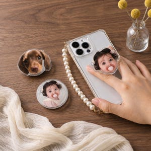 May include: A clear smartphone case with a pearl beaded wrist strap and a circular phone grip featuring a baby's portrait. Two additional circular phone grips are included: one with a baby's portrait and one with a brown dachshund puppy's face.
