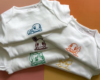 Könnte beinhalten: Eine Sammlung von fünf weißen kurzärmeligen Babybodys, die jeweils mit einer unterschiedlichen Charakter-Stickerei in blauem, braunem, orangefarbenem, grünem oder gelbem Garn versehen sind. Die Kleidungsstücke sind gestapelt und zeigen die Stickereien.