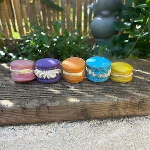 Peut inclure: Cinq macarons colorés, rose, violet, orange, bleu et jaune, chacun avec une garniture différente. Les macarons sont disposés sur une surface en bois vieilli, avec de la verdure et une statue en arrière-plan.