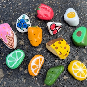 May include: A selection of painted stones, each designed to look like different foods. The stones are painted to resemble an ice cream cone, blueberries, a strawberry, a fried egg, a pepper, a slice of pizza, an avocado, a lime, an orange slice, a green pepper, and a lemon slice.