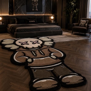 May include: A large, shaped area rug featuring a stylized cartoon character with a skull-like head, X-shaped eyes, and a brown hooded sweatshirt. The rug has a thick, textured pile with black, cream, and brown color blocking, designed in a character silhouette.