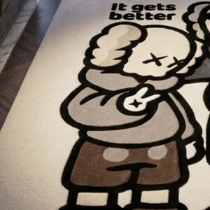 May include: A rectangular area rug featuring a cream-colored background with a large, stylized cartoon character graphic in shades of brown, gray, and black. The character has X-shaped eyes and is making a peace sign. The text "It gets better" is printed.
