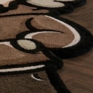 May include: A tufted rug featuring a stylized cartoon character design with thick black outlines. The rug is composed of brown, beige, and cream-colored yarn sections, creating a textured, multi-layered appearance with rounded, organic shapes.