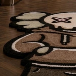 May include: A tufted area rug shaped like a cartoon character with a brown hoodie, tan face, and black outlines. The character features X-shaped eyes, white gloved hands, and a brown lower body section, all rendered in a plush, textured pile material.