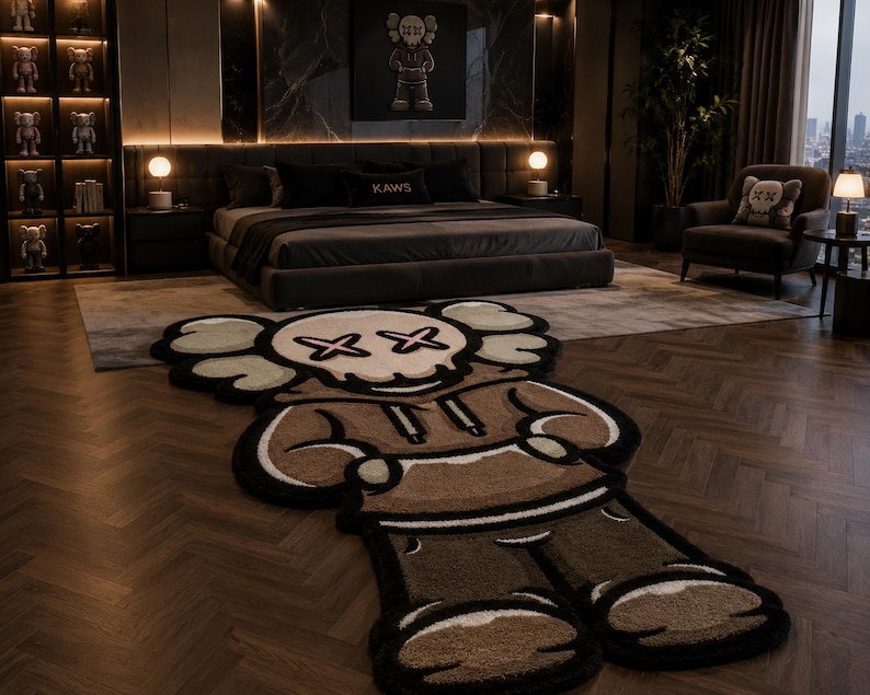 May include: A large, shaped area rug featuring a stylized cartoon character with a skull-like head, X-shaped eyes, and a brown hooded sweatshirt. The rug has a thick, textured pile with black, cream, and brown color blocking, designed in a character silhouette.