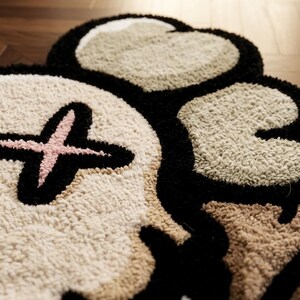 May include: A tufted rug featuring a stylized skull design with a pink cross over one eye socket. The rug is constructed with thick, textured yarn in shades of cream, beige, and black, creating a high-pile surface with defined, bold outlines.