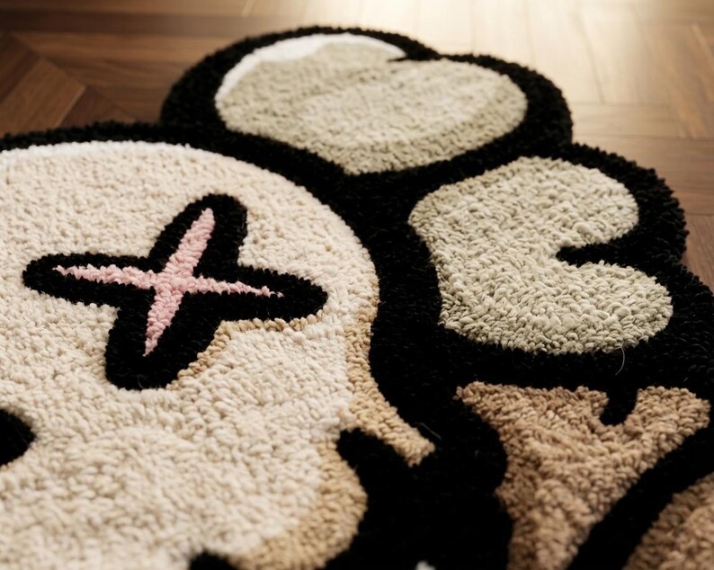 May include: A tufted rug featuring a stylized skull design with a pink cross over one eye socket. The rug is constructed with thick, textured yarn in shades of cream, beige, and black, creating a high-pile surface with defined, bold outlines.
