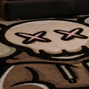 May include: A plush area rug featuring a stylized character design with a tan face, black outlines, and pink cross-shaped eyes. The rug incorporates brown, white, and light green color blocks in a cartoon-inspired graphic pattern with a soft texture.