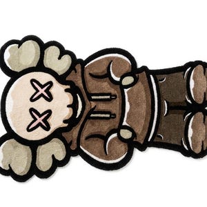 May include: A tufted rug shaped like a cartoon character with a skull-like head, cross-shaped eyes, and cloud-like ears. The figure wears a brown hooded sweatshirt and dark brown pants, outlined in thick black yarn with a soft, textured surface.