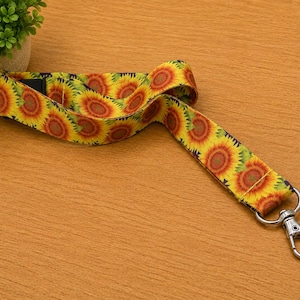 May include: A fabric lanyard featuring a repeating pattern of yellow and orange sunflowers with brown centers against a dark background. The strap includes a silver-toned metal swivel lobster clasp attached to one end for securing items.
