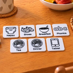 May include: A set of seven white square cards with black line art illustrations and text labels. The cards feature icons for Nappy, Shoes, Breakfast, Phonics, Tea, Dinner, and Tablet, designed as visual communication aids for daily routines.