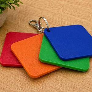 May include: A set of four square-shaped plastic tags in red, orange, green, and blue, each featuring a rounded corner design. The tags are connected together by a silver-coloured metal split ring and a lobster claw clasp, allowing for easy attachment.