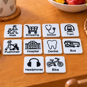 May include: Ten square white tiles featuring black line art icons and text labels for daily activities and locations: Walk, Shopping, Doctor, Train, Pushchair, Hospital, Dentist, Bus, Headphones, and Bike, arranged in a grid on a wooden surface.