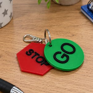 May include: A keychain featuring two flat plastic tags attached to a silver metal ring and lobster clasp. One tag is a red hexagon with the word STOP embossed in black, and the other is a green circle with the word GO embossed in black.