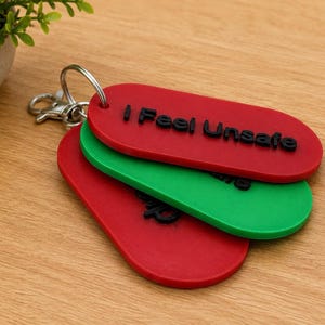 Puede incluir: Un conjunto de tres llaveros de forma ovalada unidos a una anilla de metal plateado. El llavero superior es rojo con el texto en relieve negro I Feel Unsafe. El llavero central es verde y el llavero inferior es de color rojo.