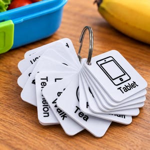 Peut inclure: Un ensemble de cartes de communication carrées blanches maintenues par un anneau métallique. Chaque carte présente un dessin au trait noir et du texte, incluant les mots Tablet, Tea et Walk. Les cartes sont disposées en éventail.