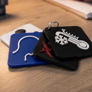 May include: A set of three square keychains with rounded corners, featuring raised icons. One black keychain displays a white thermometer and snowflake, one blue keychain shows a white outline of a head, and one white keychain has a black circular icon.