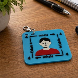 May include: A square blue keychain featuring a graphic of a person with black hair and a red shirt. The design includes black text reading Inhale and Exhale with arrows and the number 4, creating a visual guide for a box breathing exercise.