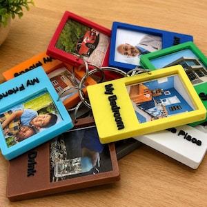 May include: A collection of rectangular plastic photo frame keychains in various colors including red, blue, green, yellow, orange, brown, and white. Each frame holds a unique photograph and features black text labels such as My Friend and My Bedroom.