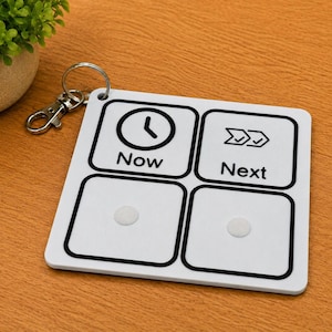 May include: A white square keychain with rounded corners featuring four black-bordered sections. The top left section shows a clock icon and the text Now, while the top right shows a double checkmark icon and the text Next. Two blank sections are below.