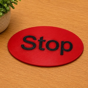 May include: An oval-shaped red coaster featuring the word Stop in raised black lettering. The surface of the coaster has a textured finish, and the letters are clearly defined against the vibrant red background of the oval object.