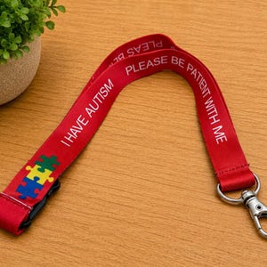 May include: A red fabric lanyard featuring the white printed text I HAVE AUTISM and PLEASE BE PATIENT WITH ME. The strap includes a colorful puzzle piece graphic in blue, yellow, and green, and is finished with a silver-toned metal lobster clasp.