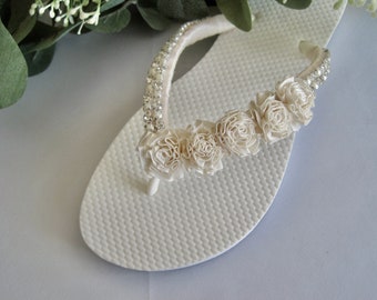 May include: White flip flops with a white satin strap adorned with rhinestones and a row of white fabric flowers.