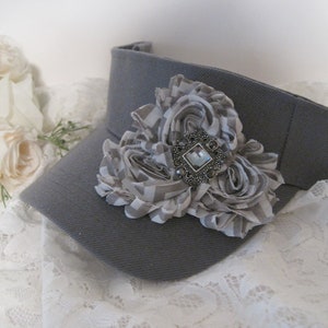 May include: A gray visor with a large gray and white fabric flower with a silver and clear rhinestone embellishment.