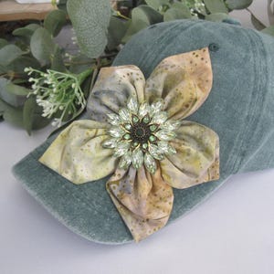 May include: A teal baseball cap adorned with a large, decorative flower. The flower is made of fabric with a watercolor-like pattern in shades of yellow, brown, and gray. The flower's center is a sparkling jewel. The cap is a casual accessory.