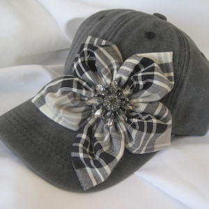 May include: A charcoal gray baseball cap featuring a large, fabric flower embellishment on the front. The flower is crafted from black, white, and gray plaid fabric and is centered with a cluster of clear, faceted crystals.