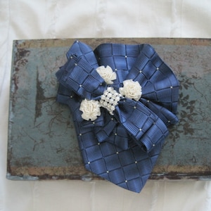 May include: A navy blue bow with white flowers and a rhinestone center. The bow is on a blue and brown textured surface.
