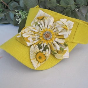 May include: A yellow visor with a large sunflower-patterned fabric flower attached to the front. The flower has a jeweled center.