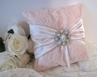 May include: A pink lace pillow with a white satin bow and a pearl and rhinestone brooch. The pillow is perfect for a wedding ceremony or reception.