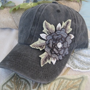 May include: A dark gray baseball cap with a large embroidered flower in shades of brown, gray, and white. The flower has a rhinestone center.
