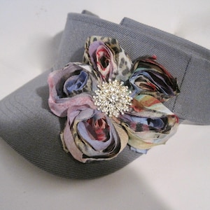 May include: A gray visor with a decorative fabric flower. The flower has a rhinestone center and petals in shades of pink, purple, blue, and animal print. The visor is made of a textured fabric.