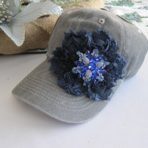 May include: A gray baseball cap with a blue fabric flower and a large blue rhinestone brooch.