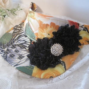 May include: A floral visor with a black lace flower and a rhinestone embellishment. The visor has a butterfly pattern and is made of a lightweight fabric.