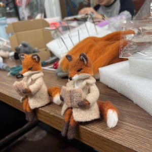 May include: Two needle-felted fox figurines with orange and white fur, each wearing a beige coat and holding a small, open book. The foxes have pointed ears, dark eyes, and dark brown paws, and are positioned in a seated pose on a wooden surface.