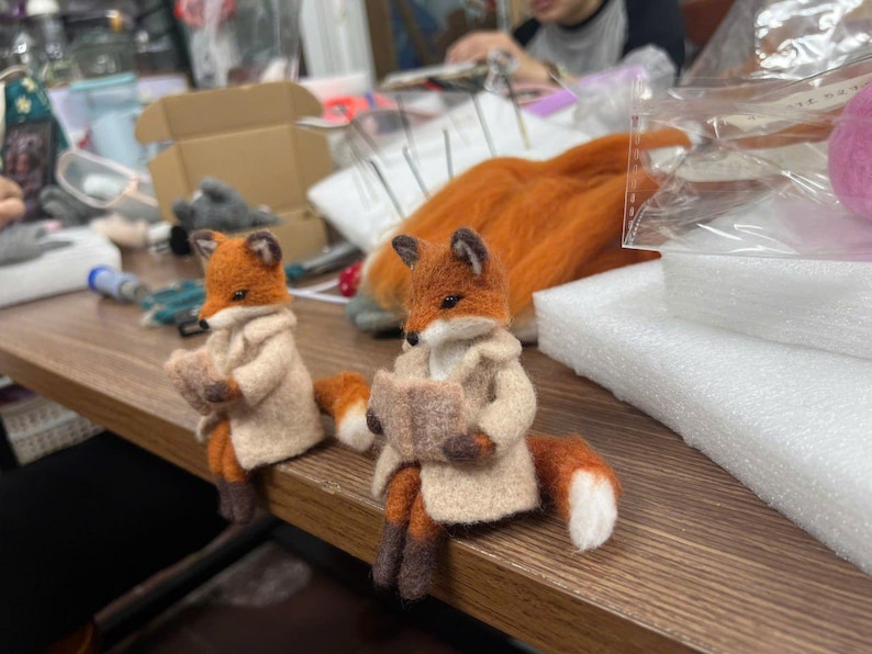 May include: Two needle-felted fox figurines with orange and white fur, each wearing a beige coat and holding a small, open book. The foxes have pointed ears, dark eyes, and dark brown paws, and are positioned in a seated pose on a wooden surface.