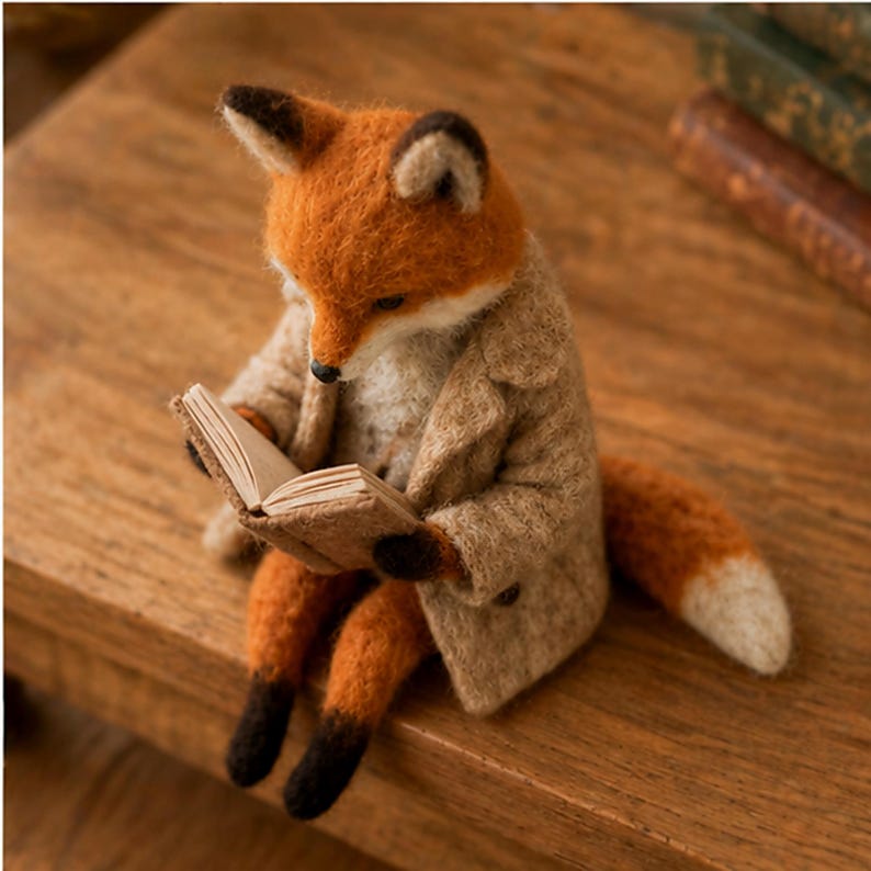 May include: A needle-felted fox figurine with orange and white fur, wearing a textured beige coat. The fox is sitting upright and holding a small, open book with light-colored pages between its paws, looking down at the text with a focused posture.