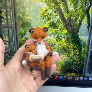 May include: A needle-felted sculpture of an adult fox and a small kit sitting together. The adult fox has orange fur with a white chest and dark brown paws, holding a small, open, light-colored book. The kit is nestled against the adult.