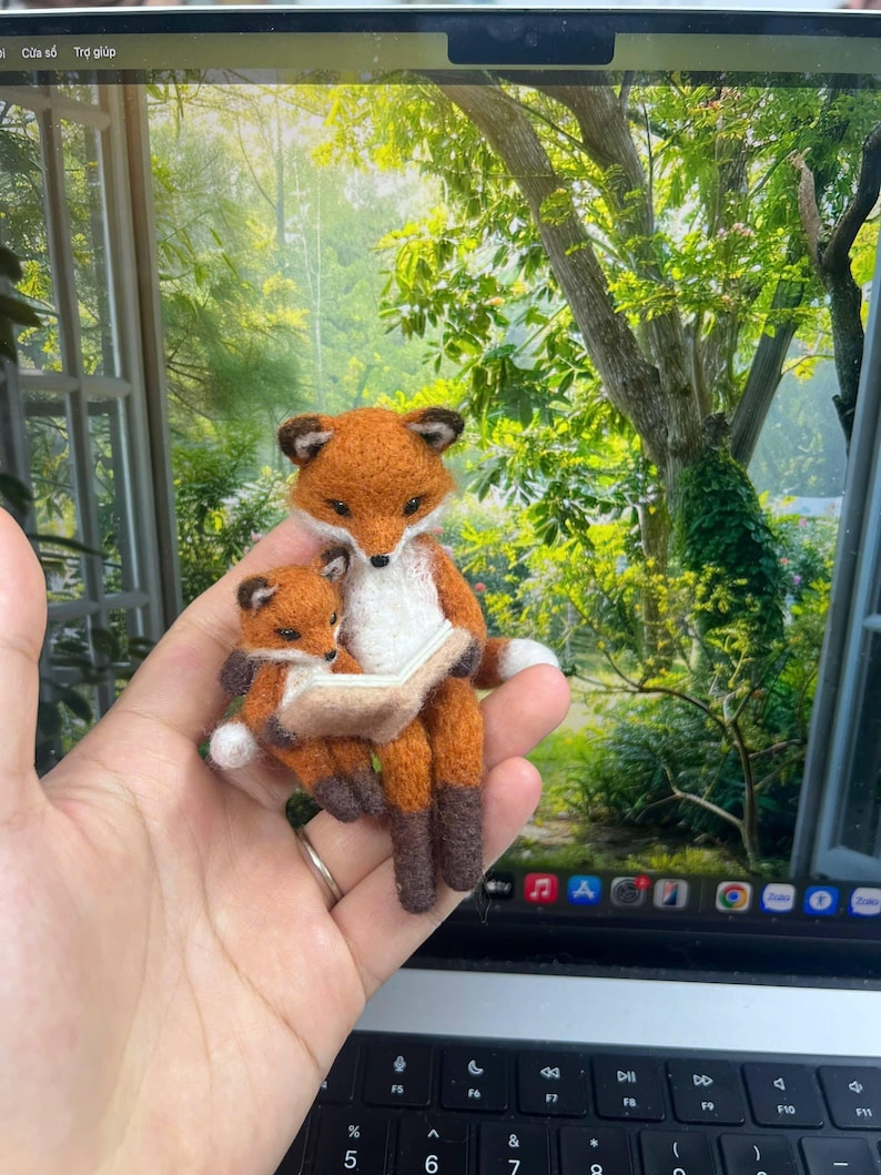May include: A needle-felted sculpture of an adult fox and a small kit sitting together. The adult fox has orange fur with a white chest and dark brown paws, holding a small, open, light-colored book. The kit is nestled against the adult.