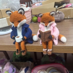 May include: Two needle-felted fox figurines sitting side-by-side. The left fox wears a navy blue coat and holds a small white mug. The right fox wears a light pink coat and holds a small brown book. Both foxes have orange fur, white chests, and brown shoes.