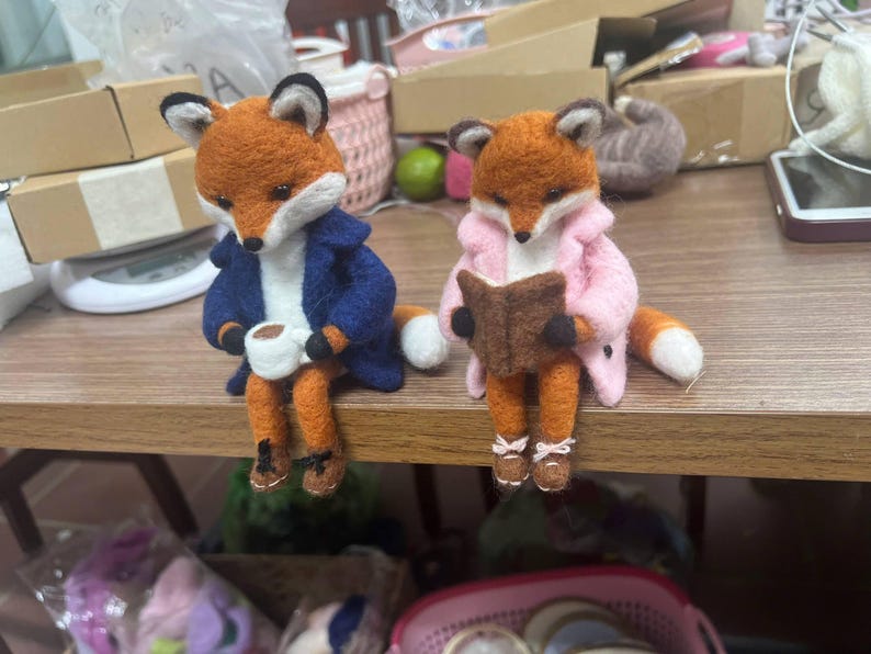 May include: Two needle-felted fox figurines sitting side-by-side. The left fox wears a navy blue coat and holds a small white mug. The right fox wears a light pink coat and holds a small brown book. Both foxes have orange fur, white chests, and brown shoes.